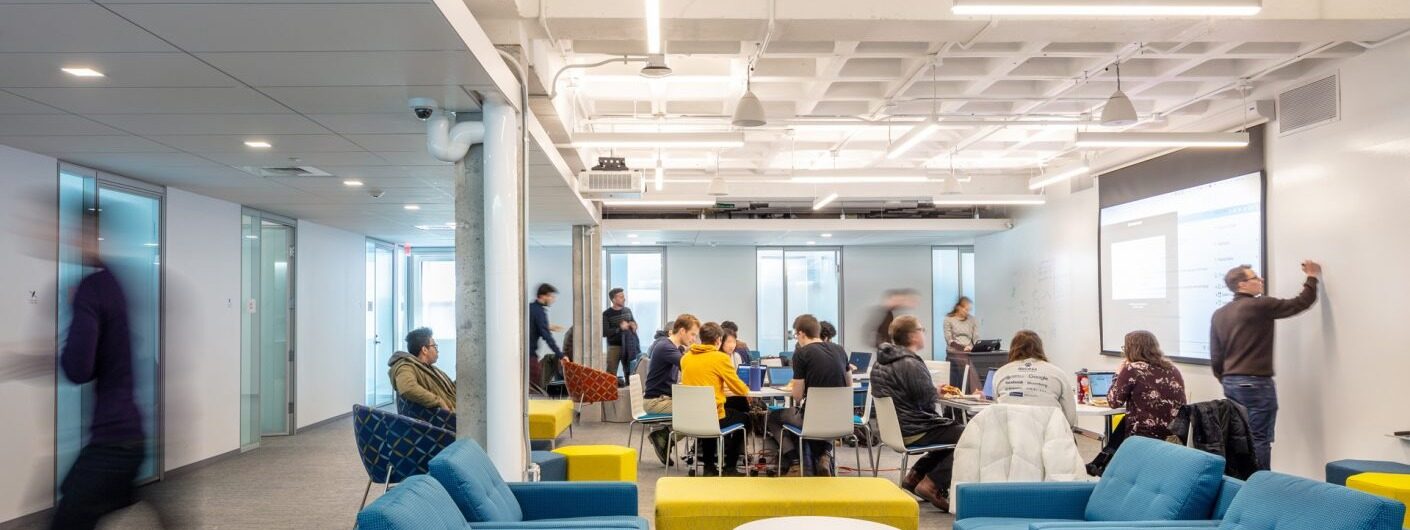 The Carney Institute innovation hub: an open floor with students working together at tables with laptops, someone writing on a whiteboard beside a large screen, and glass-walled meeting rooms along one side